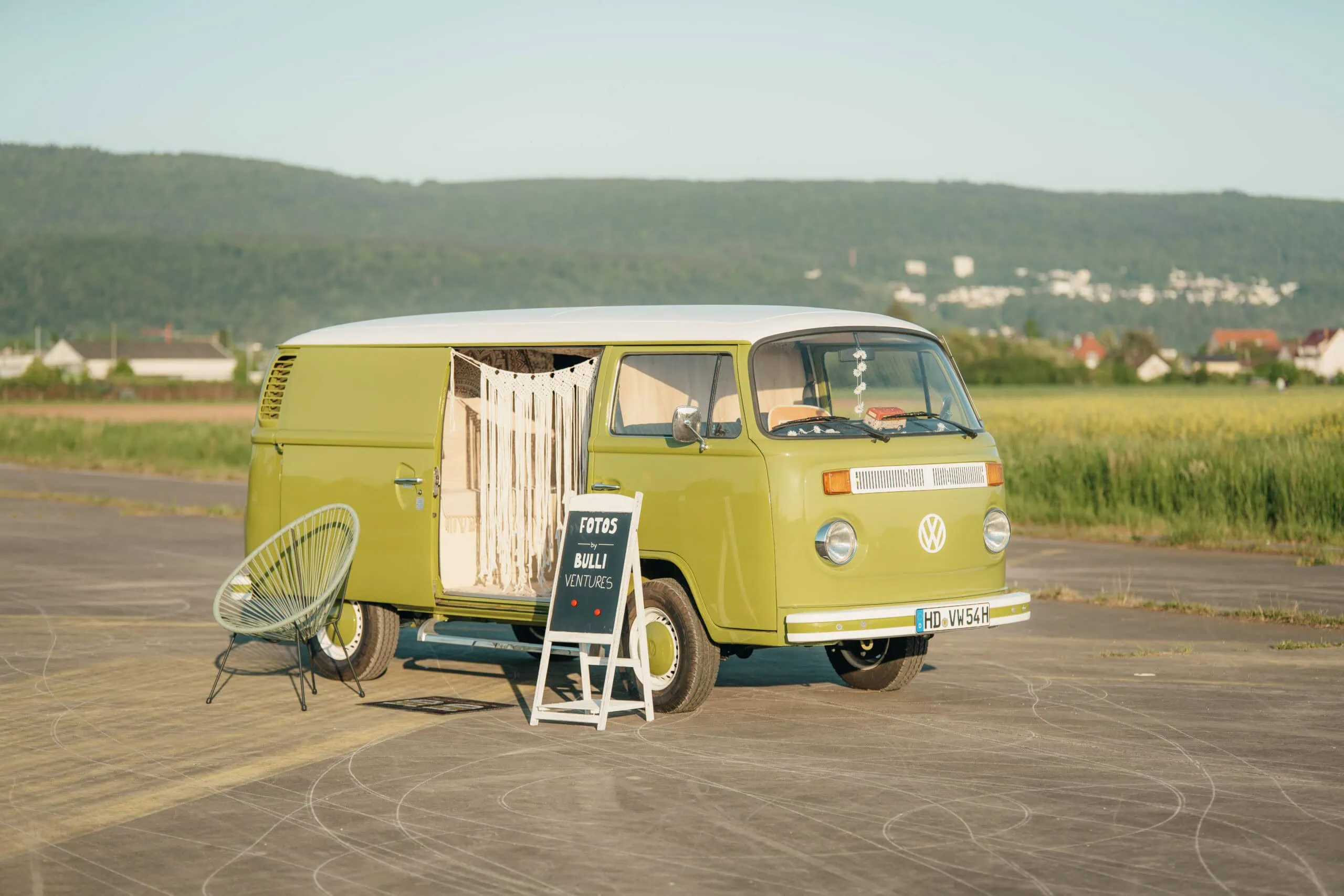 Grüner VW T2 Fotobulli Martha am Straßenrand, daneben ein Gartenstuhl und ein Schild.