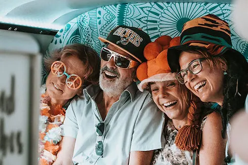 Vier Erwachsene mit bunten Hüten, Sonnenbrillen und Kostümen posieren im Fotobus bei einer Firmenfeier.