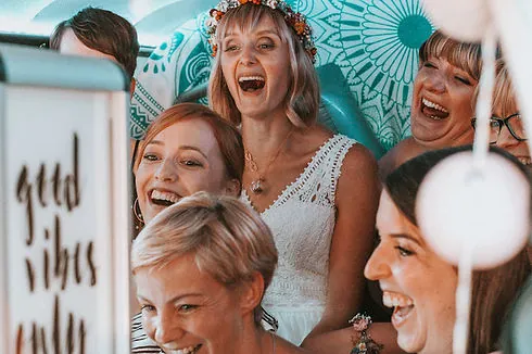 Hochzeitsgäste lachen gemeinsam im Fotobulli - die Fotobox im VW T2 ist mit gemusterten Stoffen und Lichterketten dekoriert.