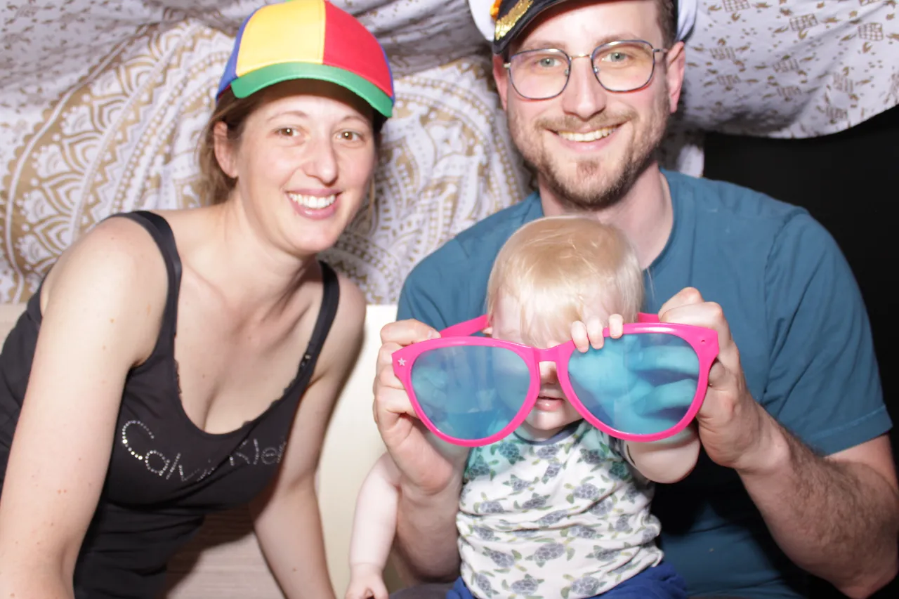 Familie im Fotobulli: Mama mit bunter Cap, Papa mit Kapitänsmütze und Baby mit großer pinker Spaß-Sonnenbrille.